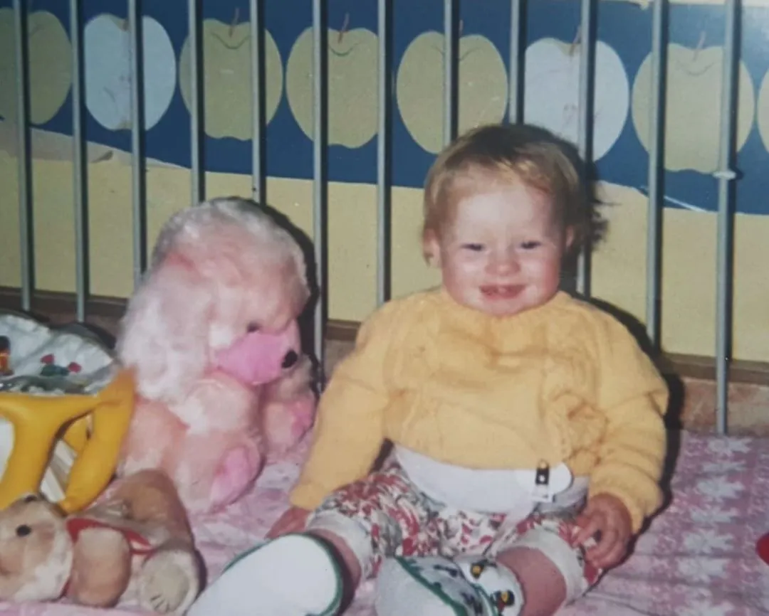 A picture of Danielle as a toddler, sat in a hospital crib surrounded with stuffed animals. She's smiling widely.